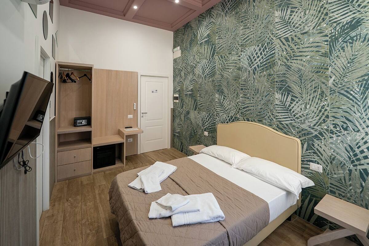 A hotel room with a bed, a TV, a wardrobe, a desk, and a wall with a leaf pattern.
