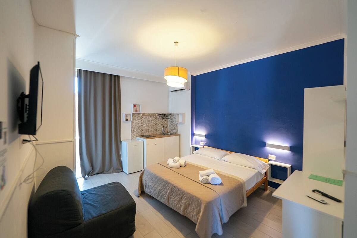 A hotel room with a bed, bedside table, desk, and a kitchenette.