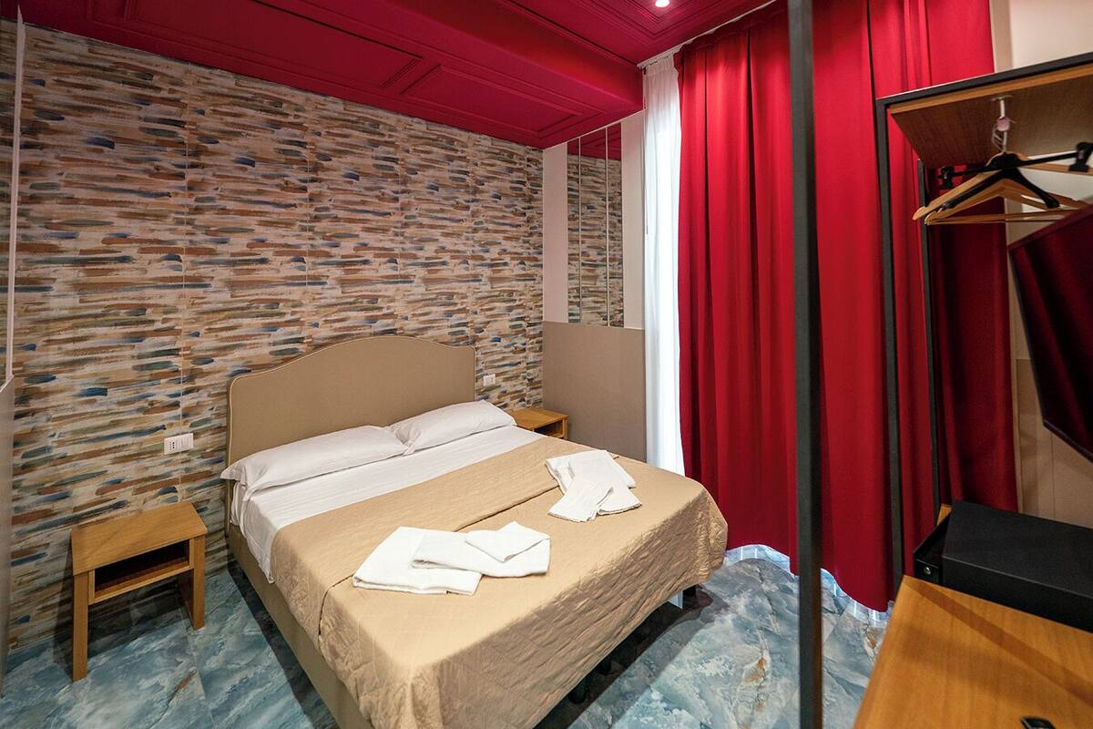 A hotel room with a stone-patterned wall, a bed with a beige cover, a wooden nightstand, and a red curtain.