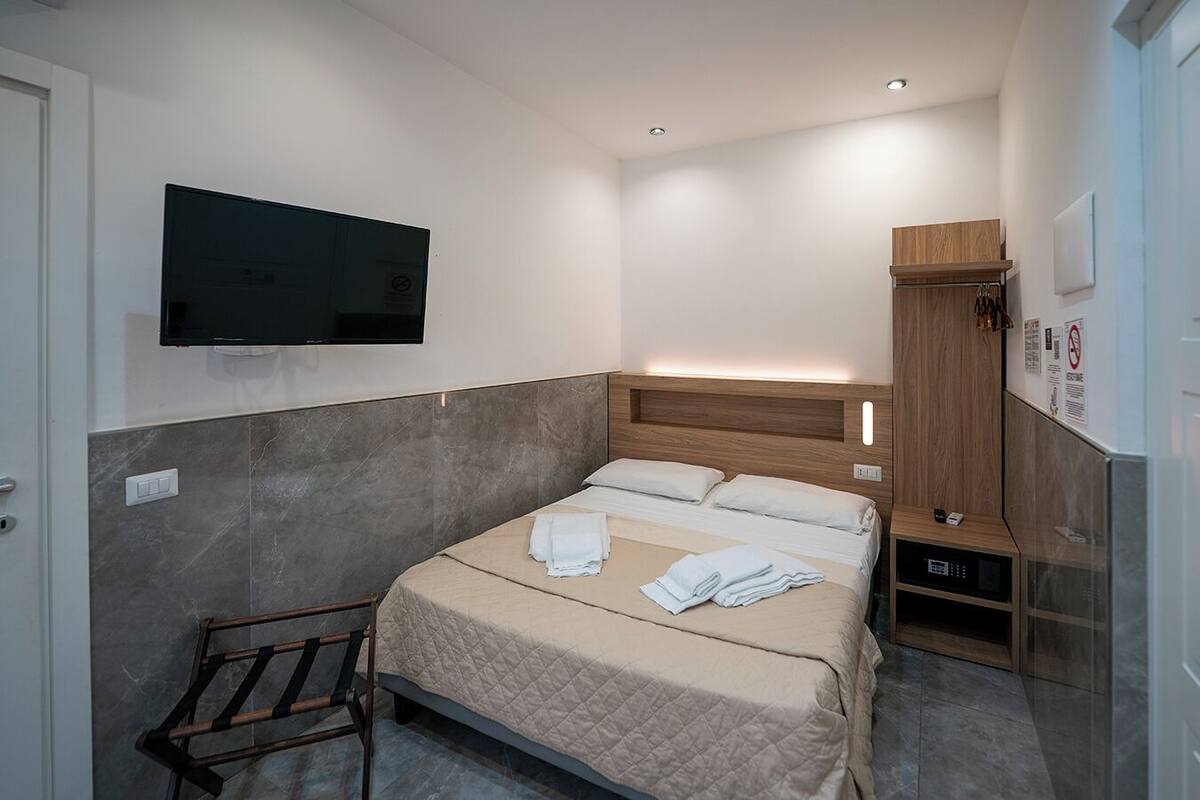 A modern hotel room with a bed, a TV mounted on the wall, a wooden headboard, a bench, and a microwave.