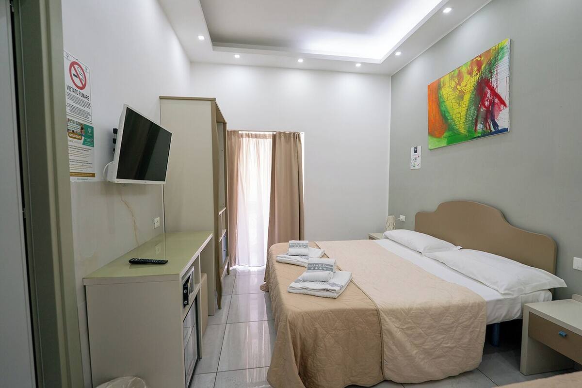 A hotel room with a bed, bedside tables, a desk with a TV, a painting on the wall, and a bathroom visible through a door.