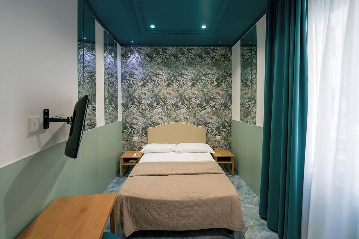 A narrow hotel room with a bed, a small table, and a wall-mounted TV.