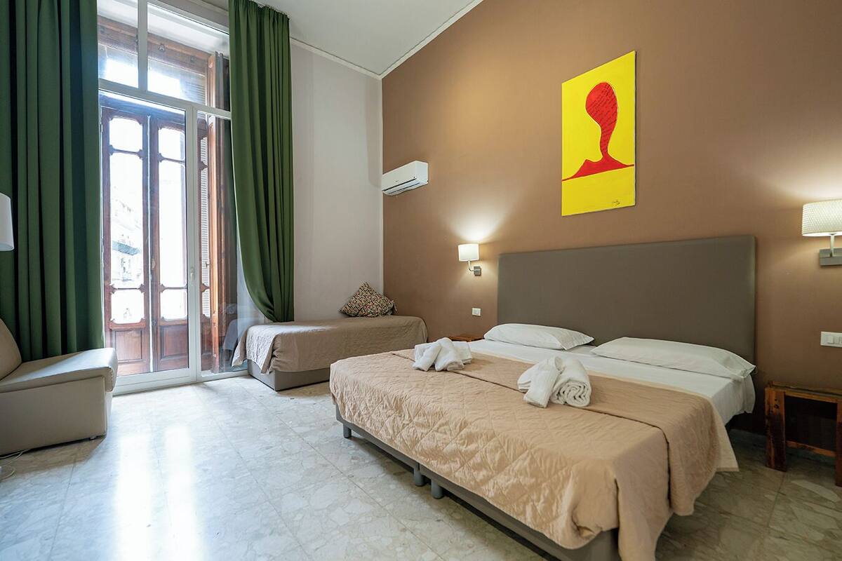 A hotel room with two beds, a sofa, a large window with curtains, a wall-mounted air conditioner, and a painting.
