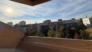 Vista desde un balcón con suelo de madera, que da a un paisaje urbano con varios edificios y cielo despejado.