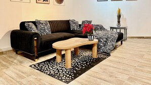 Un salon moderne avec un canapé aux motifs noir et blanc, une table basse en bois et un tapis aux motifs noir et blanc. Trois œuvres d’art encadrées ornent le mur, et une petite table est agrémentée d’un vase et d’une plante.