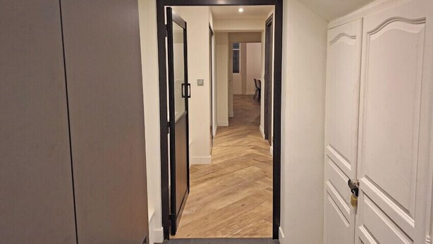 Un pasillo con piso de madera, paredes blancas y una puerta con marco negro que da a otra habitación.