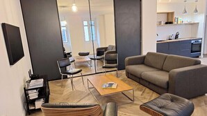 Una sala de estar moderna con sofá, mesa de centro y sillas. Se observa un área de cocina al fondo con mesa y sillas para comer.