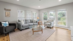 Ein geräumiges Wohnzimmer mit einem grauen Sofa, einem runden Glastisch und einem gemusterten Sessel.