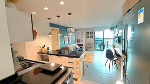 A modern kitchen with a central island, a dining area with a table and chairs, and a large refrigerator. - Aloha Residence (Ipojuca)