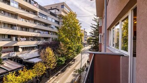 A multi-story building with balconies, a street with parked cars, and trees with autumn foliage.