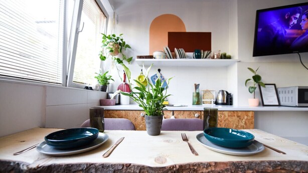 A modern kitchen with a dining table set for two, a TV mounted on the wall, and various kitchen appliances and decor.