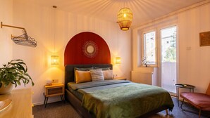 A bedroom with a bed, bedside tables, a hanging light fixture, a potted plant, and a window with blinds.