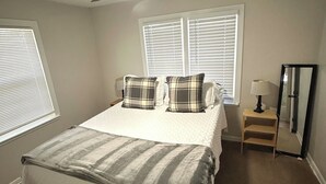 A bedroom with a bed, two windows with blinds, a ceiling fan, a bedside table with a lamp, and a mirror.