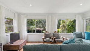 Un salon moderne doté d’une grande fenêtre, d’un téléviseur à écran plat posé sur un meuble en bois, d’un canapé vert et de deux fauteuils en bois.