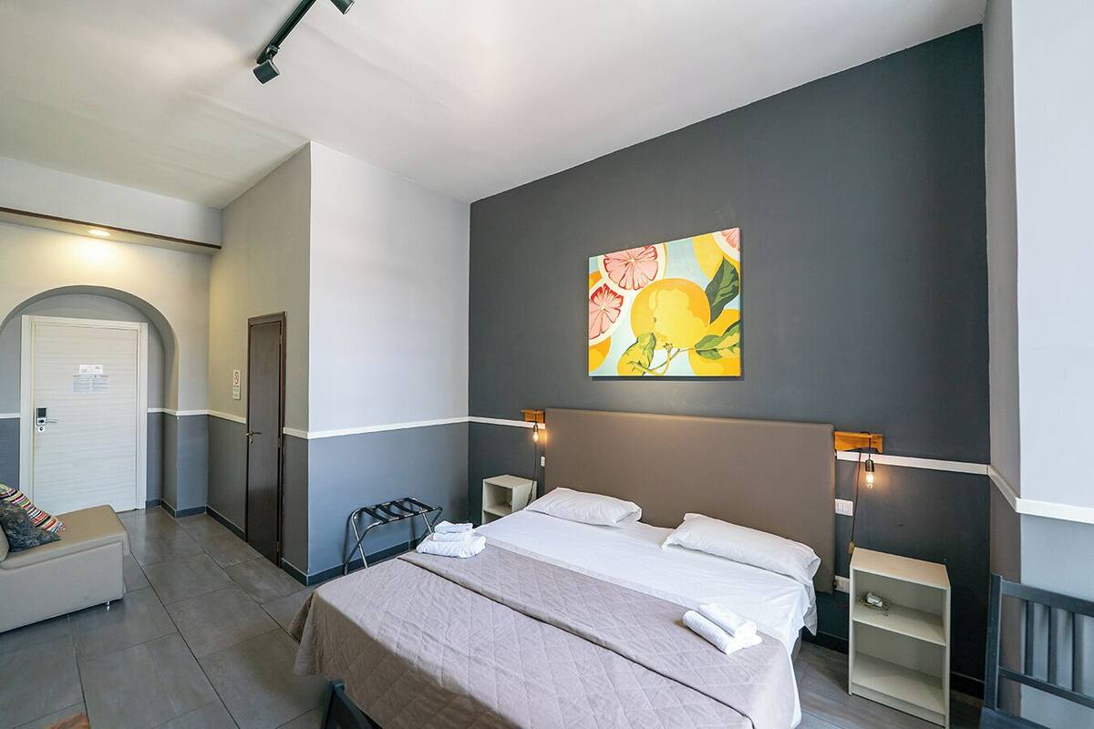 A hotel room with a bed, a seating area with a chair, a towel rack, and a painting on the wall.
