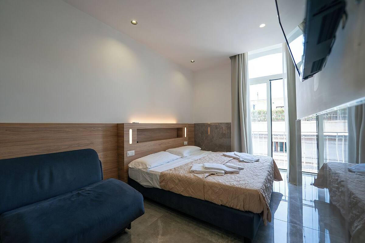 A modern hotel room with a bed, a blue sofa, and a TV mounted on the wall.