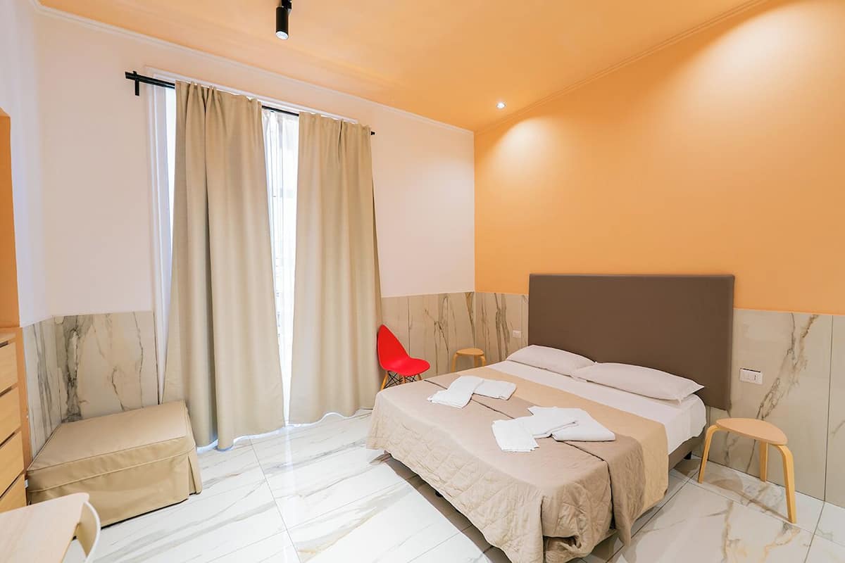 A hotel room with a bed, a chair, a small stool, a desk, and a window with curtains.
