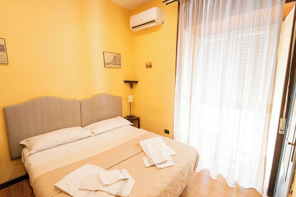 A hotel room with a bed, a nightstand, a lamp, and an air conditioning unit.