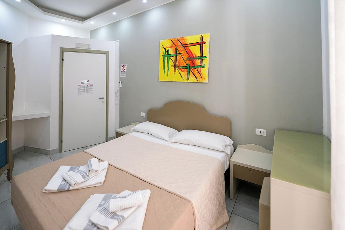 A hotel room with a bed, a bedside table, a nightstand, a door, and a painting on the wall.