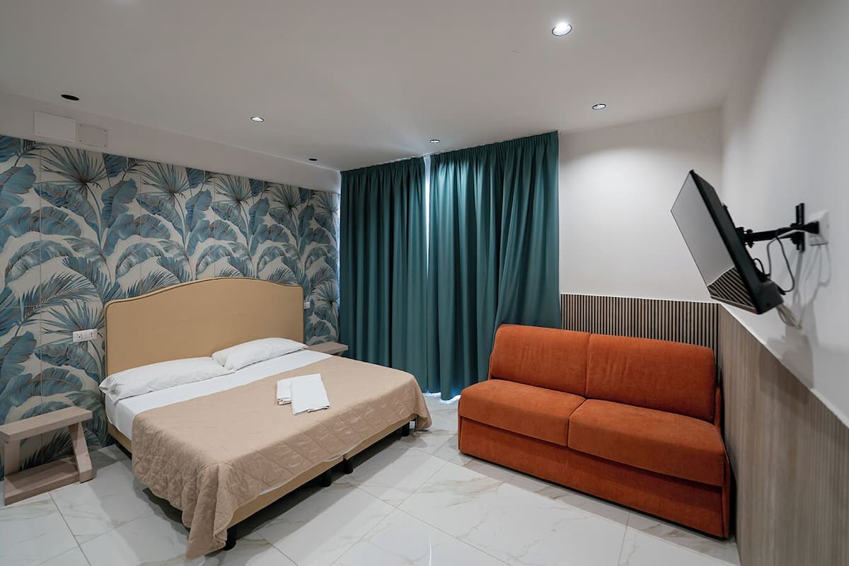 A hotel room with a bed, a sofa, a TV mounted on the wall, and a window with curtains.
