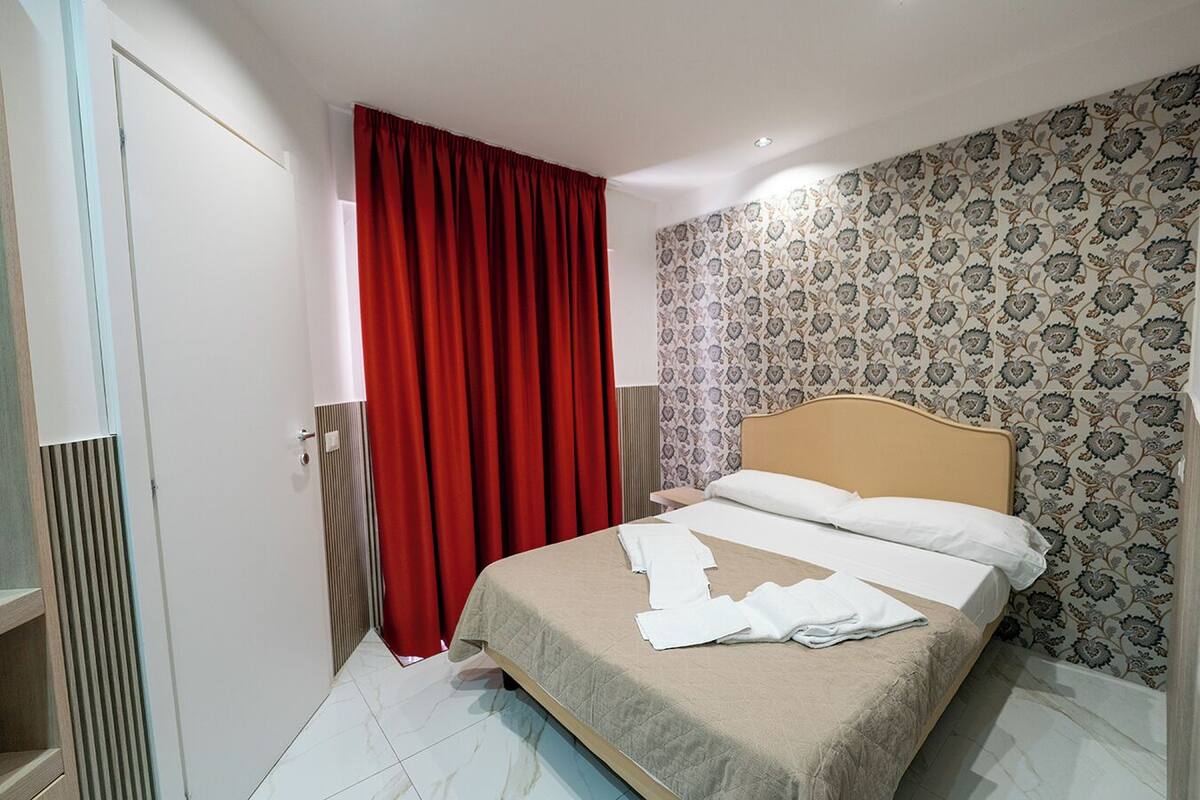 A bedroom with a bed, a red curtain, patterned wallpaper, and a radiator.