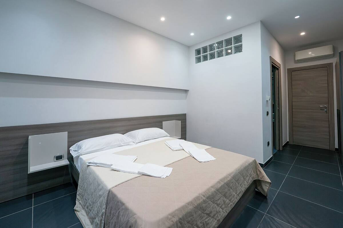 A modern hotel room with a large bed, a bedside table, a wall-mounted air conditioner, and a door with a wooden panel.
