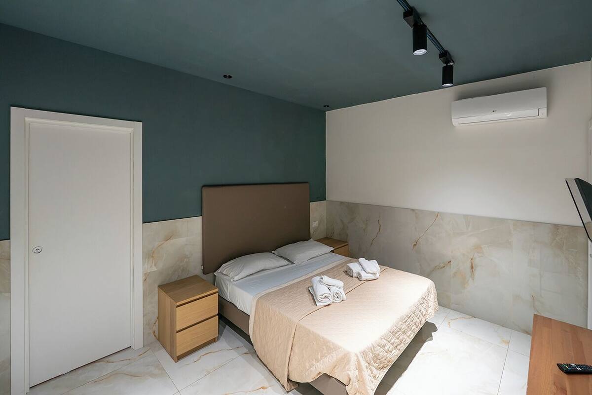 A bedroom with a bed, a nightstand, a drawer, an air conditioning unit, and a door.