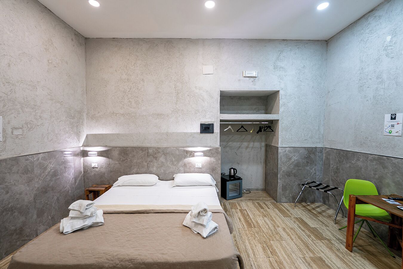 Une chambre d’hôtel compacte comprenant un lit, une petite table, une chaise et une étagère intégrée avec des crochets.