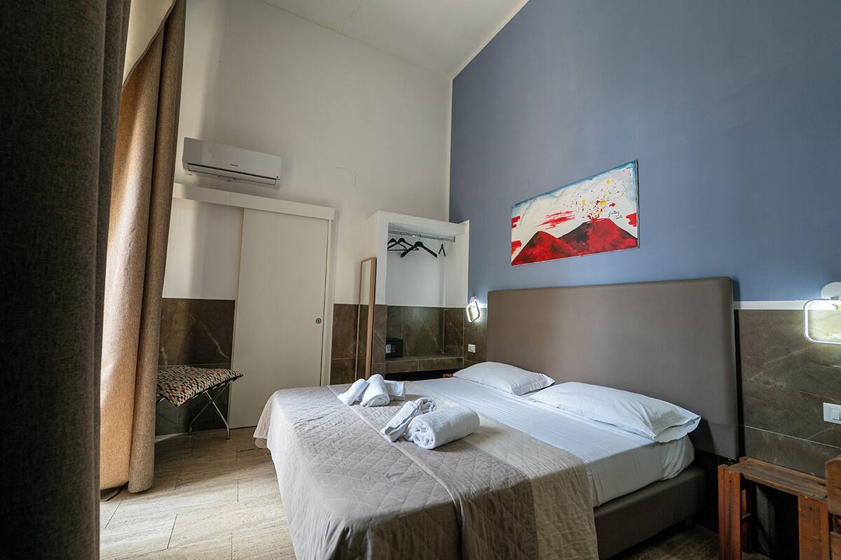 A hotel room with a bed, a bedside table, a mirror, an air conditioning unit, and a painting on the wall.