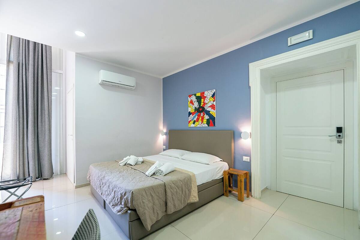 A modern hotel room with a bed, a small wooden stool, a door with a lock, and a wall-mounted air conditioning unit.
