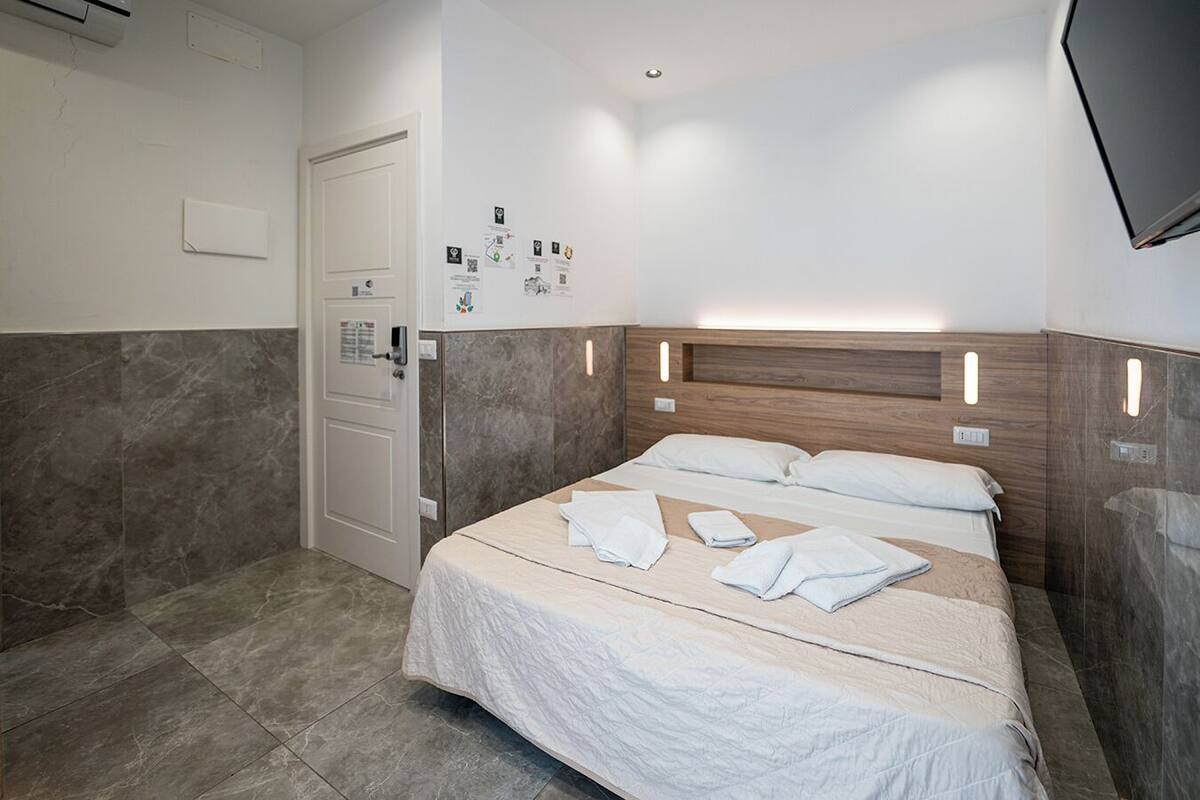 A hotel room with a bed, a door, a wall-mounted control panel, and a flat-screen TV.