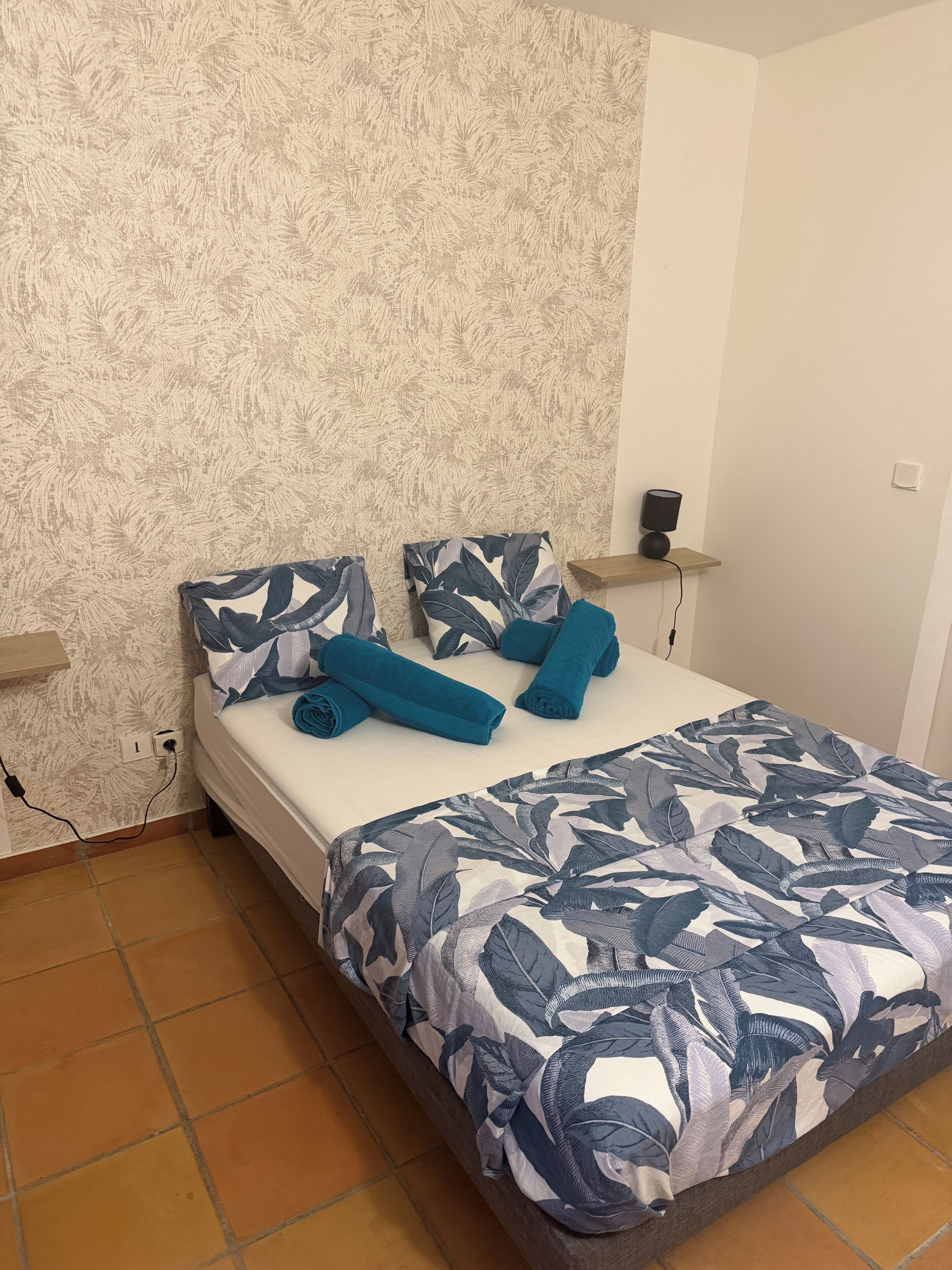 A neatly made bed with a leaf-patterned bedspread and matching pillows, a bedside lamp, and a power outlet on the wall.