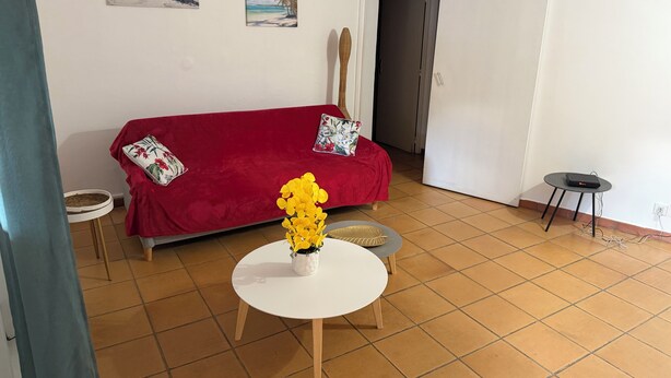 A living room with a red sofa, a white table with a yellow flower arrangement, and two beach-themed paintings on the wall. - Bungalow Korail (Pointe-Noire)