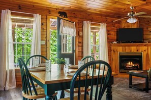 A wooden cabin interior with a dining table set for four, a fireplace, a television, and large windows with curtains. - Cabin with Hot Tub.15 min to Gatlinburg&Pigeon Forge. (Sevierville)