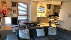 A modern living room with a dark grey sectional sofa, two white fluffy pillows, a dining table with chairs, and a kitchen in the background. - Appartement 78 m² 1 chambre, terrasse et garage (Aix-les-Bains)