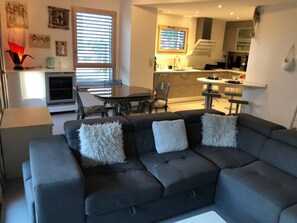 Ein modernes Wohnzimmer mit einem dunkelgrauen Ecksofa, zwei weißen, flauschigen Kissen, einem Esstisch mit Stühlen und einer Küche im Hintergrund.
