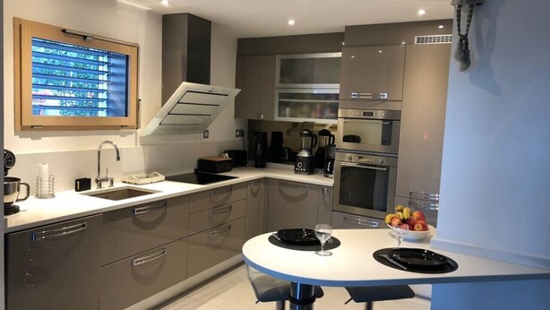 A modern kitchen with a built-in oven, a sink, and a dining area with a round table. - Appartement 78 m² 1 chambre, terrasse et garage (Aix-les-Bains)