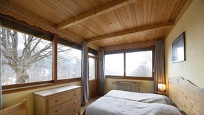 Une chambre avec un plafond en bois, un lit, une commode, une fenêtre donnant sur des arbres et un radiateur.