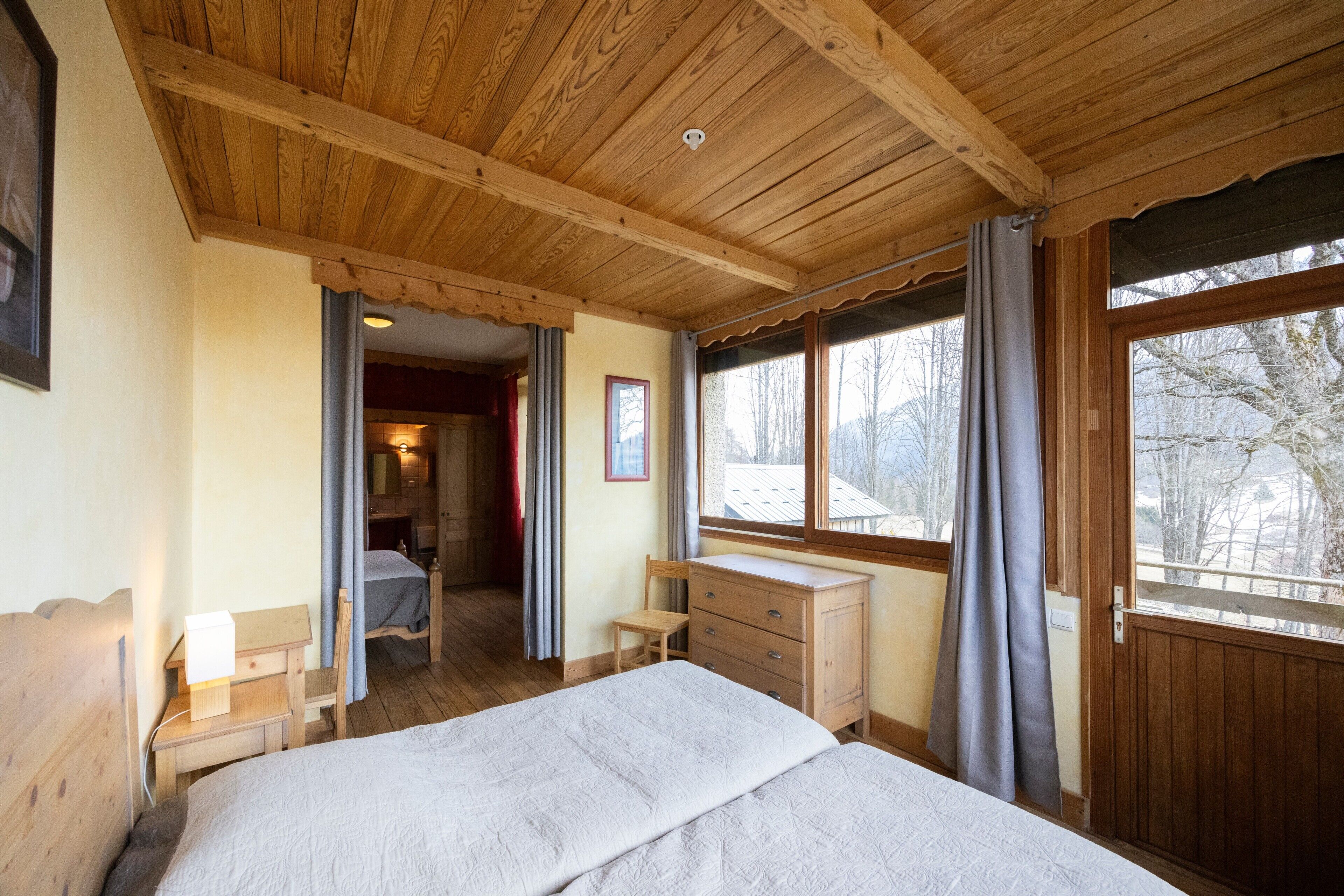 Eine Holzbalkendecke, ein Bett, ein Schminktisch, ein Stuhl und ein Fenster mit Blick auf Bäume.