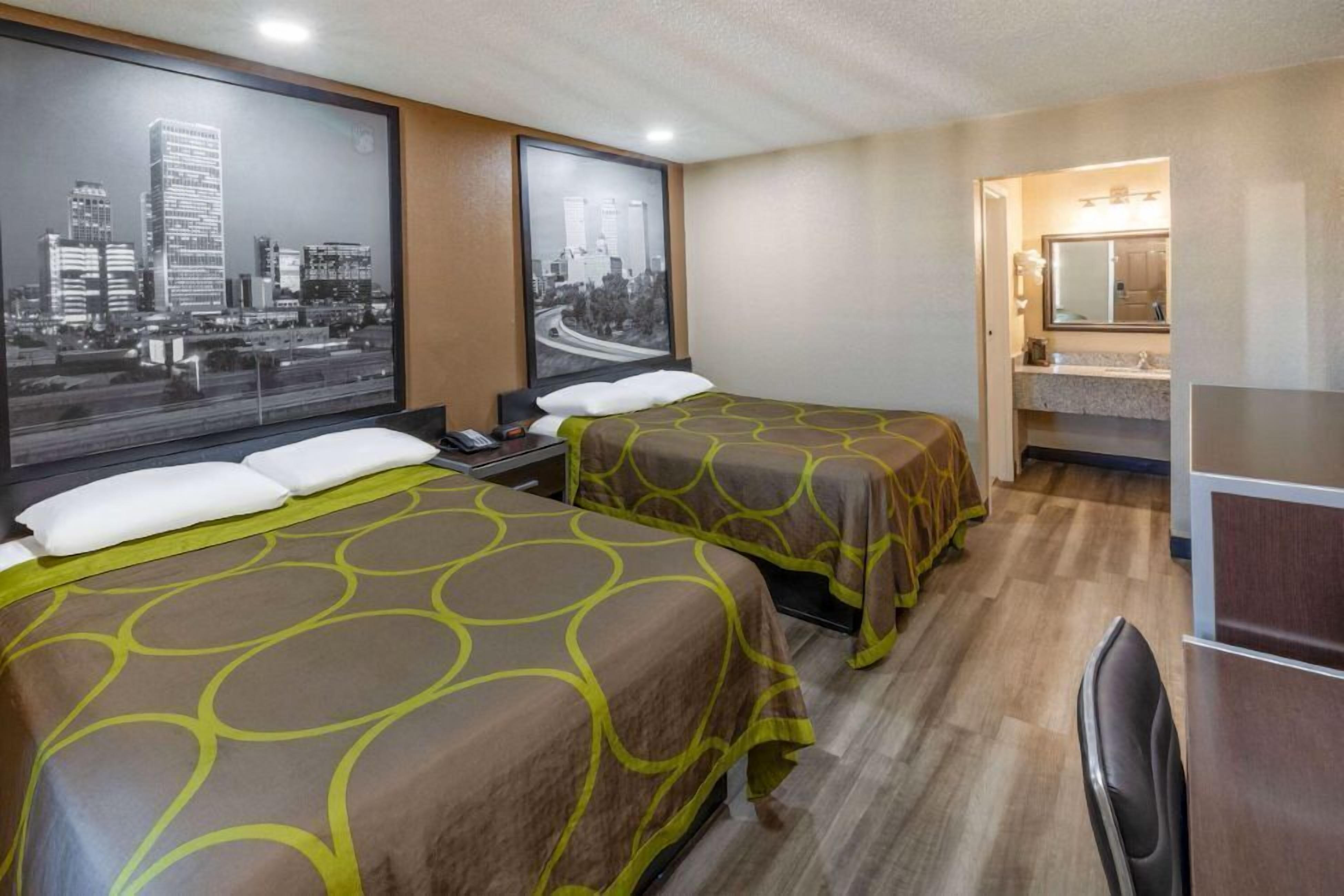A hotel room with two beds, a large window with cityscape views, a bathroom with a mirror and sink, and a desk with a chair.
