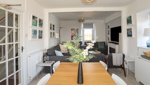 A dining area with a wooden table, white chairs, and a vase of flowers. There is a grey sofa and a television in the background. - Angorfa Apartment (Pwllheli)