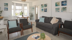 A modern living room with a grey sofa, two armchairs, a coffee table, and a dining area with a white table and chairs. - Angorfa Apartment (Pwllheli)