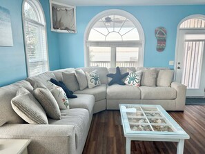 A living room with a beige sectional sofa, a glass-top coffee table, and a view through a large arched window. - Sandy Sound- Half house, BoatDock, 2 min walk to beach, Hot tub, Sleeps 8, Views (Emerald Isle)