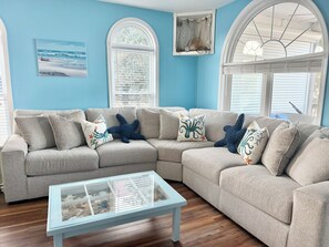 A living room with a beige sectional sofa, a glass coffee table, and a framed seascape painting. - Sandy Sound- Half house, BoatDock, 2 min walk to beach, Hot tub, Sleeps 8, Views (Emerald Isle)