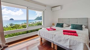 Une chambre dotée d’une grande fenêtre donnant sur la mer et une île lointaine. La chambre comprend un lit avec une literie blanche et deux oreillers, un vert et un rouge.