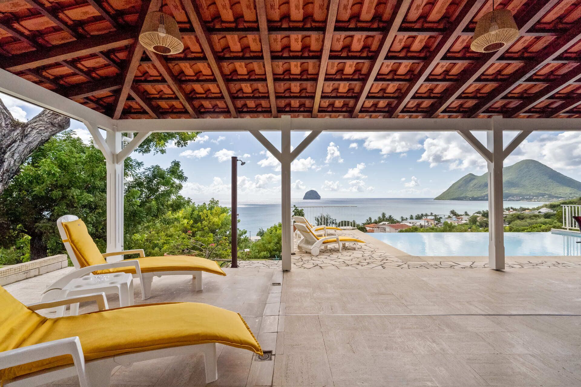 A covered outdoor area with lounge chairs and a pool, overlooking a scenic view of the sea and a mountain.
