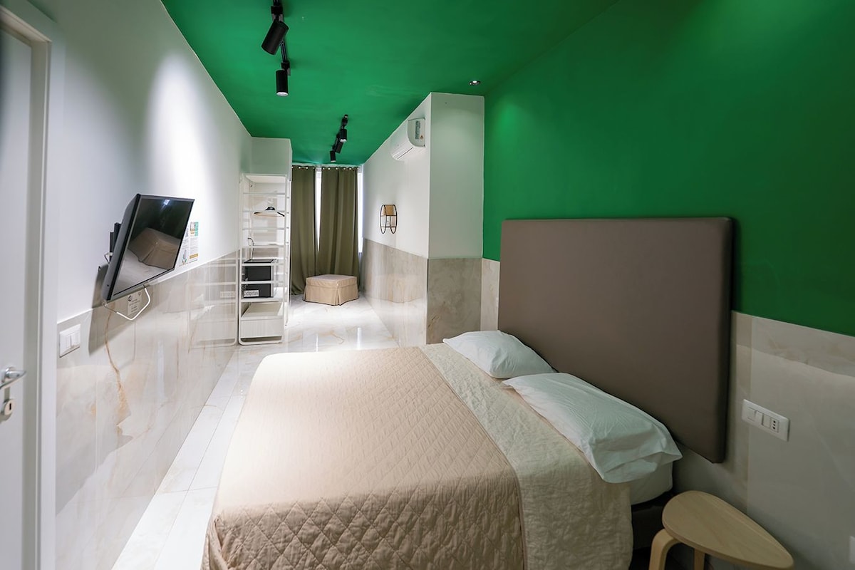 Ein Hotelzimmer mit einem Bett, einem kleinen Hocker, einem an der Wand befestigten Fernseher, einem Regal mit Gegenständen und einer grünen Akzentwand.