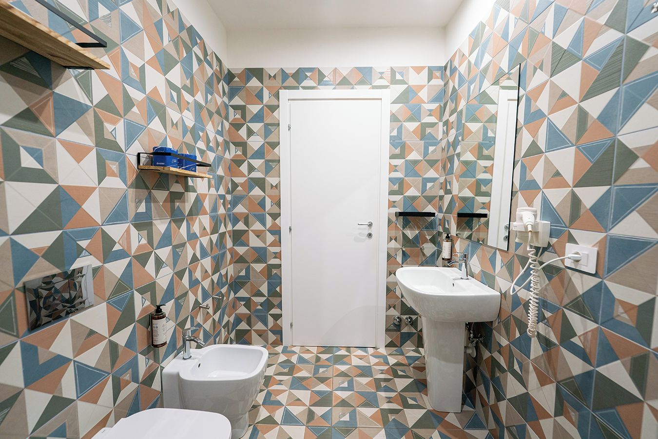 Un baño pequeño con baldosas de diseño geométrico, una puerta blanca, dos lavamanos y un bidé.