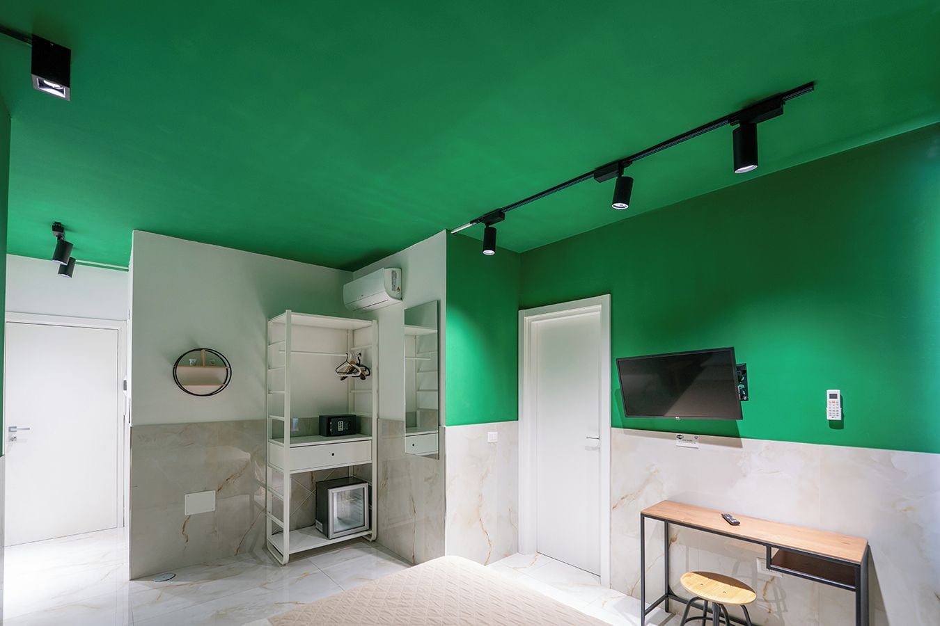 A room with a green wall, a white door, a TV mounted on the wall, a small table with a chair, and a bunk bed with a mirror.