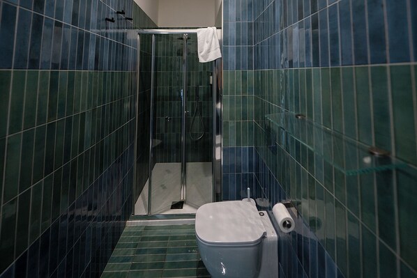 Une salle de bain avec des murs recouverts de carreaux verts, une douche en verre, des toilettes blanches et une serviette accrochée à la porte de la douche.